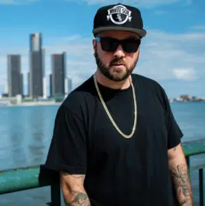 A man wearing a black t-shirt, gold chain, sunglasses, and a White Sox cap stands by a railing with a city skyline and water in the background.